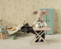 A Maileg Dad Mouse, dressed in the Maileg Wedding suit and sporting a smart bow tie, carefully irons tiny pants—perhaps preparing for a miniature wedding—in a cozy, vintage-inspired room furnished with a small bed, green cupboard, and breakfast treats on the bedside table; an ideal setting for fans of charming Maileg mouse accessories.