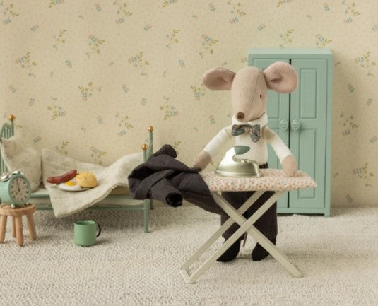 A Maileg Dad Mouse, dressed in the Maileg Wedding suit and sporting a smart bow tie, carefully irons tiny pants—perhaps preparing for a miniature wedding—in a cozy, vintage-inspired room furnished with a small bed, green cupboard, and breakfast treats on the bedside table; an ideal setting for fans of charming Maileg mouse accessories.