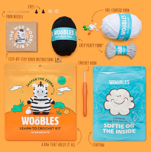 A flat lay of The Woobles Jasper the Zebra Crochet Kit—featuring yarn, crochet hook, stuffing, pre-started zebra character, video instructions, and zebra-themed packaging—neatly arranged on an orange background.