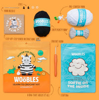 A flat lay of The Woobles Jasper the Zebra Crochet Kit—featuring yarn, crochet hook, stuffing, pre-started zebra character, video instructions, and zebra-themed packaging—neatly arranged on an orange background.