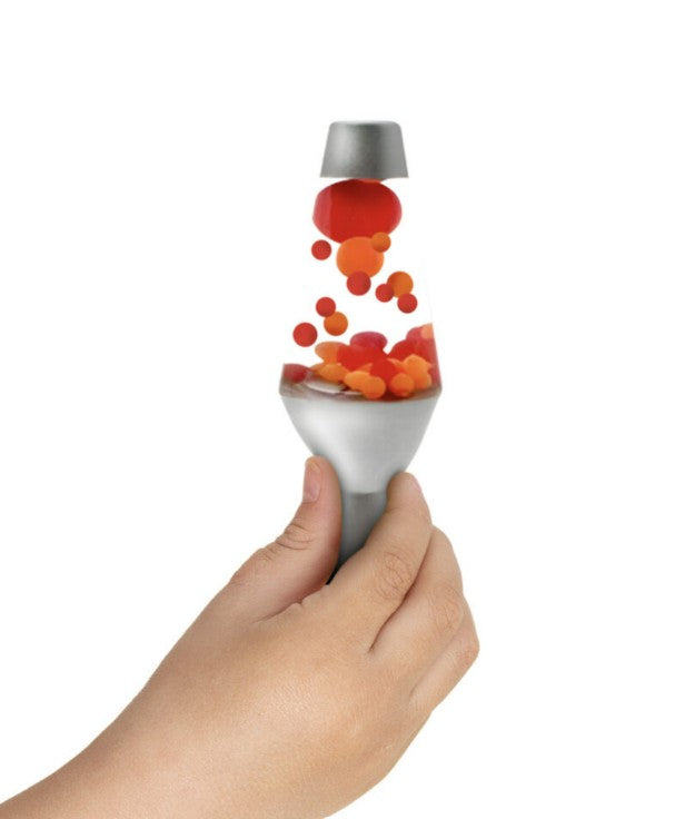 A hand is holding the Schylling Toys Lava Squish n Flow, a small silver sensory lamp with red and orange wax blobs floating inside, isolated on a white background—a mesmerizing stress relief toy.