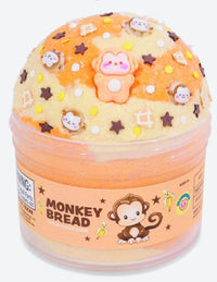 A round container labeled "Monkey Bread Slime" from Peachybbies Slime holds orange and yellow swirled sensory slime, topped with star and monkey decorations. The label features a cartoon monkey and bananas, making this Peachybbies Slime product perfect for stress relief and fun play.