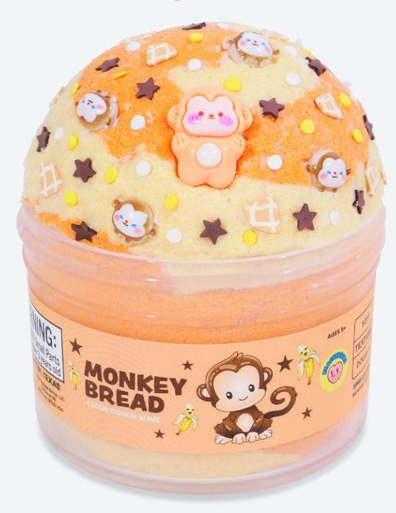 A round container labeled "Monkey Bread Slime" from Peachybbies Slime holds orange and yellow swirled sensory slime, topped with star and monkey decorations. The label features a cartoon monkey and bananas, making this Peachybbies Slime product perfect for stress relief and fun play.