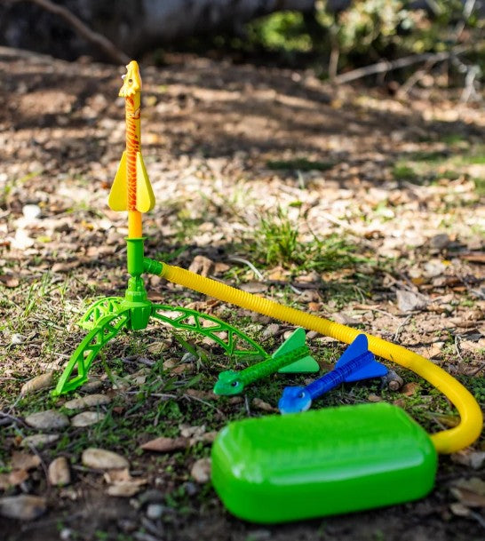 A D&L Stomprocket Dino-Soar Stomp Rocket Kit sits on the ground, equipped with a yellow hose, green foot pump, and vibrant dinosaur-themed rockets—ideal for outdoor play among grass and rocks while sparking STEM exploration.