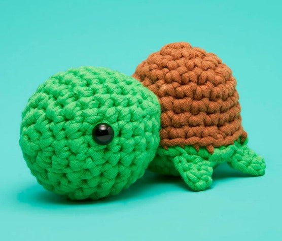 Emilio the Turtle Crochet Kit by The Woobles features a small crochet turtle with a green head and legs, brown shell, and black bead eye on a turquoise background—a delightful choice for beginner crafters.