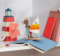 A Maileg Beach Mice, Surfer Big Brother by Maileg rests on a miniature sailboat atop an open blue book. Behind it, a red lighthouse, a doll’s sweater, a red book, and colorful shells are arranged on a white shelf—an ideal setup for any beach toy enthusiast or fan of the Surfer Big Brother mouse.