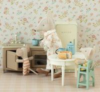Three toy mice enjoy a vintage-style kitchen with dollhouse furniture and floral wallpaper. One cooks, another opens the oven, while the third sits at a table with bread and the Maileg High Chair, Mouse - Mint by Maileg nearby.