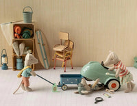 Three Maileg mice use the Maileg Wagon, Mouse - Blue as they pretend to repair a tiny toy car; one mouse lifts the car, another works underneath, and the third looks on. In the background are shelves filled with camping gear, chairs, and a surfboard. The playful scene from Maileg encourages cozy and imaginative pretend play with their miniature blue wagon.