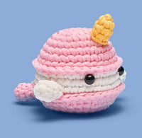 The Woobles’ Bjorn the Narwhal: Sweetie Edition Crochet Kit lets beginners craft a cute pink and white narwhal plush with a yellow horn, black button eyes, and small tail, all set against a light blue background.
