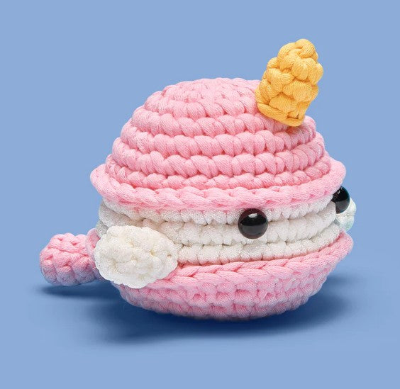 The Woobles’ Bjorn the Narwhal: Sweetie Edition Crochet Kit lets beginners craft a cute pink and white narwhal plush with a yellow horn, black button eyes, and small tail, all set against a light blue background.