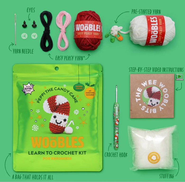 A flat lay of The Woobles Peppi the Candy Cane Crochet Kit with yarn, crochet hook, stuffing, yarn needle, safety eyes, video instructions, and a festive green bag—perfect for beginner crochet projects.