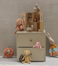 Children’s toys and stuffed animals are displayed on and around a small, pale dresser with two drawers. On top sits the Maileg SuitCasa, featuring little sister and brother mouse by Maileg, along with a dollhouse and bottle, while plush animals are nestled on the drawers and scattered across the floor—creating an inviting space for imaginative play.