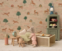 The Maileg Little Sister Mouse In Matchbox by Maileg rests in her matchbox bed with pillows and a blanket, surrounded by stools, a clock, a cup, treats, and wallpaper with horses, trees, and rainbows.