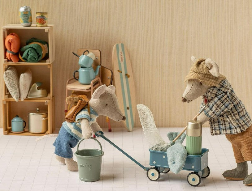 Two Maileg mice, each clad in charming outfits, get ready for an adventure. One mouse pushes the Maileg Wagon, Mouse - Blue by Maileg—its miniature frame filled with supplies—while the other carries a small bucket. Behind them, shelves lined with mini camping gear create a cozy and playful backdrop, perfect for inspiring imaginative pretend play.