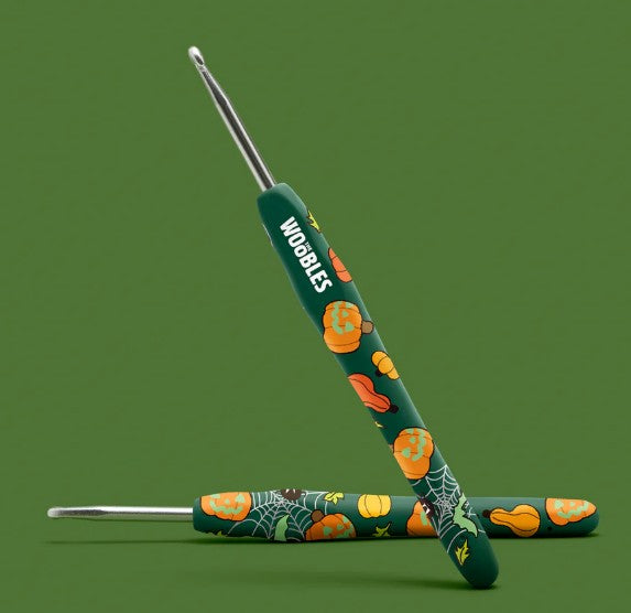 Two green crochet hooks from The Woobles’ Hal the Jack-o-Lantern Crochet Kit, each with festive jack-o’-lantern, gourd, and spider web handle designs—perfect for adding Halloween flair to your beginner crochet kit. Set on a plain green background.