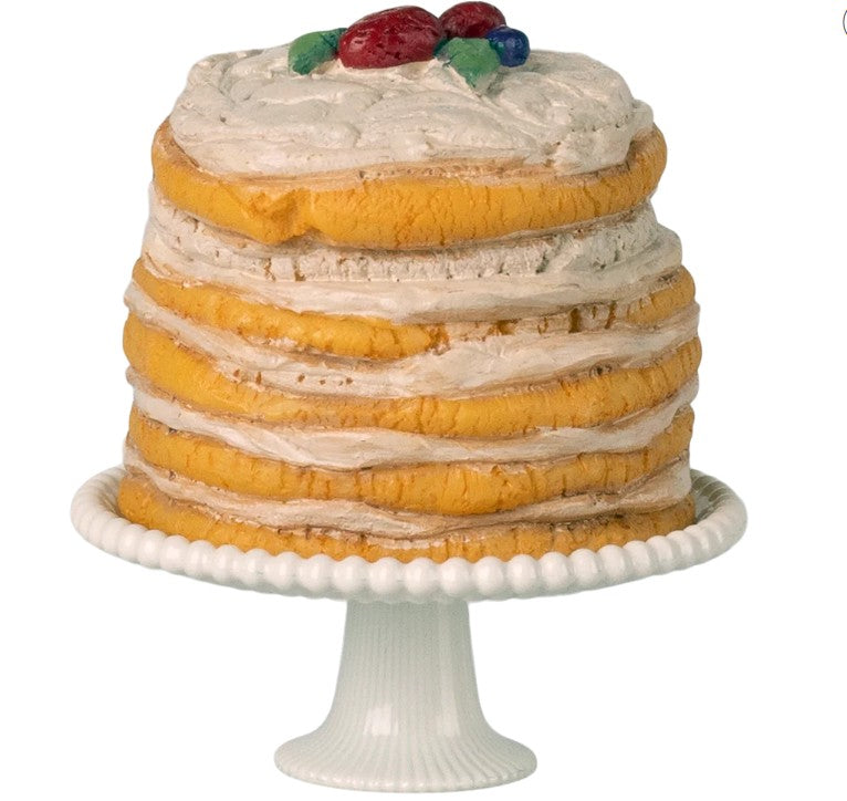The Maileg Party Cake And Cake Stand, Mouse - Off White by Maileg features a layered sponge cake with white frosting and colorful fruit on top, set on a white pedestal stand—ideal for any dollhouse party.