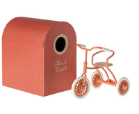 A small orange tricycle is parked beside the Maileg Tricycle Garage (Coral), labeled "Abri à Tricycle," with a round front opening—ideal for storing Maileg tricycles or Maileg Mouse accessories.