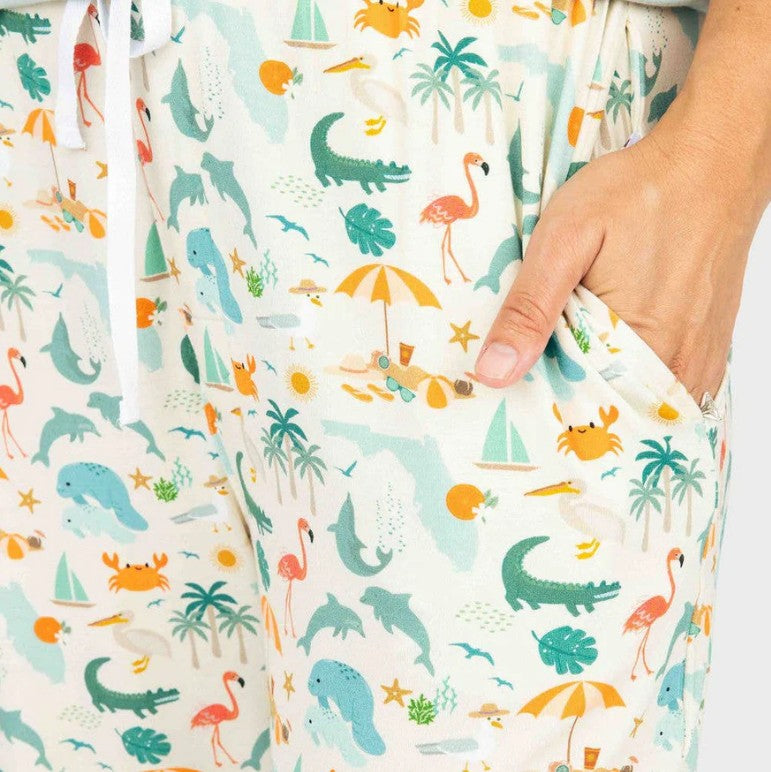 Close-up of Emerson & Friends Explore Florida Souvenir Bamboo Adult Pajamas Lounge Pants in Beige/Multi (size S), with a hand in the pocket, showcasing a vibrant tropical print featuring dolphins, flamingos, alligators, palm trees, crabs, and beach items.
