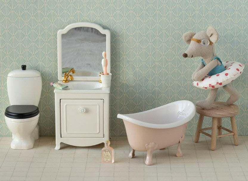 A playful bathroom scene featuring the Maileg Bathtub, Mouse - Rose by Maileg, a toilet, sink with mirror, and a stuffed mouse in glasses and swim ring on a stool. The backdrop showcases light blue patterned wallpaper.