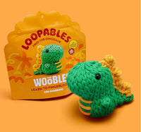 A green and yellow crocheted dinosaur toy sits before its orange box labeled "Fred the Dino Loopables Kit" by The Woobles—an ideal fingerloop craft project for beginners.