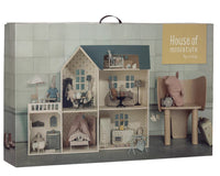 A Maileg House of miniature - Dollhouse from Maileg, complete with tiny furniture and small animal dolls, is beautifully presented in its box and beckons you into a whimsical world of make-believe. To the right of the Maileg dollhouse, a large chair adorned with a cuddly stuffed animal adds to the inviting, cozy atmosphere.