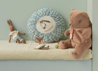A plush teddy bear with a pink scarf sits beside a brown bunny doll, a round blue plaid cushion, and Maileg’s Beach Mouse With Float, Little Brother - Yellow Dots on a cream bench against a light green wall.