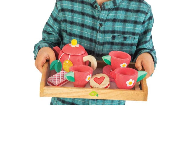 A child in a blue plaid shirt holds the Tenderleaf Tea Tray Set, featuring a red teapot, three pink cups with saucers, a sugar bowl, and a toy cookie—perfect for imaginative play.