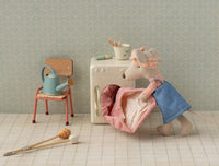 The Maileg Hiker Mouse, Big Sister, dressed in a blue outfit, loads a quilt into a toy washing machine. A nearby chair with wooden tools and watering can invite outdoor play—all with charming patterned wallpaper as a backdrop.