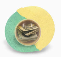 The Mystery Real Money Bath Bomb Surprise by Purposeful Bliss - Fizzy Magic is a round, green and yellow bath bomb with a clear center displaying folded U.S. dollar bills and a silver chain inside.