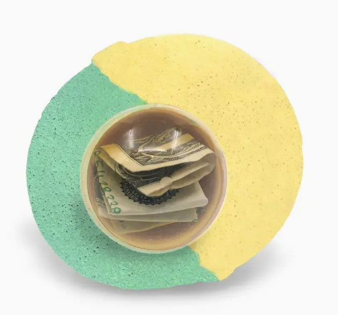 The Mystery Real Money Bath Bomb Surprise by Purposeful Bliss - Fizzy Magic is a round, green and yellow bath bomb with a clear center displaying folded U.S. dollar bills and a silver chain inside.