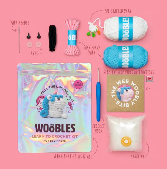 Flat lay of The Woobles Billy the Unicorn Crochet Kit, featuring yarn, eyes, needle, hook, stuffing, instructions, and a holographic bag—all designed for beginners with a fun unicorn theme.