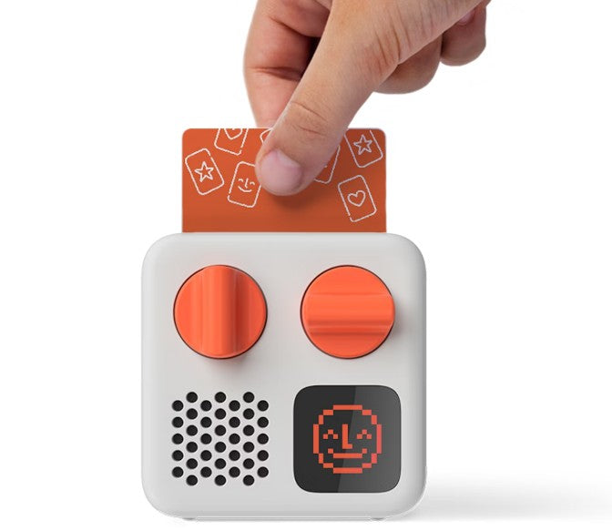 A hand inserts an orange card with symbols into the Yoto Mini (2024 Edition) by Yoto—a compact white kids’ audio player with two orange knobs, a speaker, and a digital screen showing a pixelated smiling face.