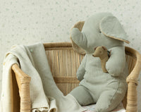 A soft fabric elephant toy sits in a wicker chair, holding the Maileg Lullaby Friends Mouse Rattle in Light Umber. A pale knitted blanket drapes over the chair, with gentle patterned wallpaper as a backdrop. Brand: Maileg.