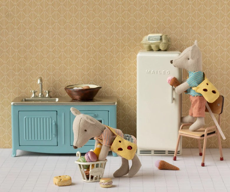 Two stuffed mice with cheese-shaped backpacks play in the Maileg Kitchen, Mouse - Blue by Maileg. One stands on dollhouse furniture eating ice cream, while the other picks up treats from the floor near a blue cabinet.