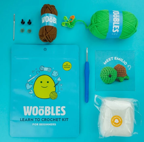The Woobles Emilio the Turtle Crochet Kit for beginners includes yarn, hook, needles, safety eyes, stuffing, a blue pouch, and a Meet Emilio card with sea turtle instructions—perfectly arranged on a blue background.