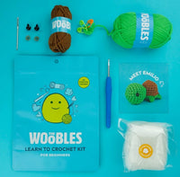The Woobles Emilio the Turtle Crochet Kit for beginners includes yarn, hook, needles, safety eyes, stuffing, a blue pouch, and a Meet Emilio card with sea turtle instructions—perfectly arranged on a blue background.