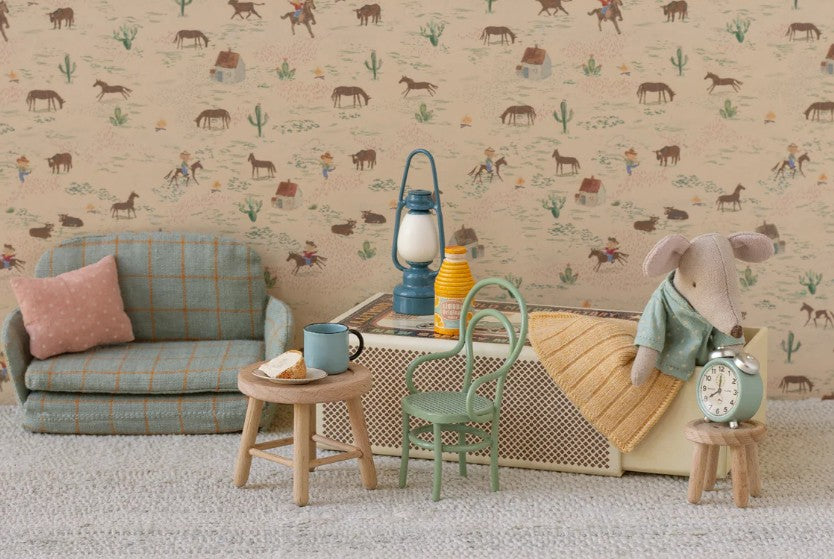 The Maileg Little Brother Mouse In Matchbox by Maileg rests on his matchbox bed in a miniature room with Western-themed wallpaper, pastel furnishings, a sofa, table with tea and cake, chairs, a clock, and a lantern.