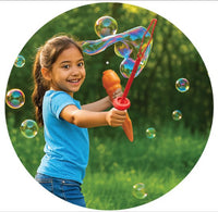 A young girl in a blue shirt smiles while making giant bubbles outdoors with the South Beach Bubbles Unspillables Bubble Warriorz, standing on green grass with trees behind her as several bubbles float around for carefree play.