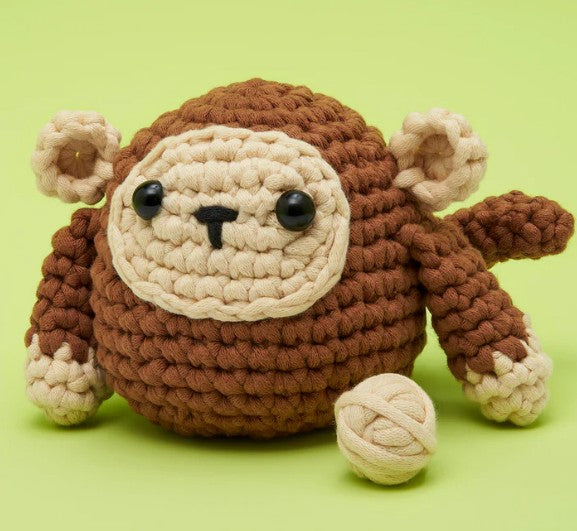 A round, crocheted brown and beige monkey made with The Woobles Jack the Monkey Crochet Kit sits on a light green surface beside a small beige yarn ball.