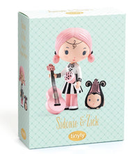 Box featuring the DJECO Tinyly Sidonie & Zick figurine, with Sidonie—a doll with pink hair holding a pink guitar—accompanied by her small black and pink pet, Zick. The packaging displays the names "Sidonie & Zick" and "tinyly" on a light patterned background, making this adorable miniature collectible by DJECO perfect for fans of charming figure sets.