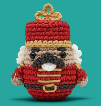 Hazel the Nutcracker Crochet Kit by The Woobles features a red hat and uniform, gold accents, black mustache, white curly hair, and black button eyes—perfect for step-by-step video lessons, set against a teal background.