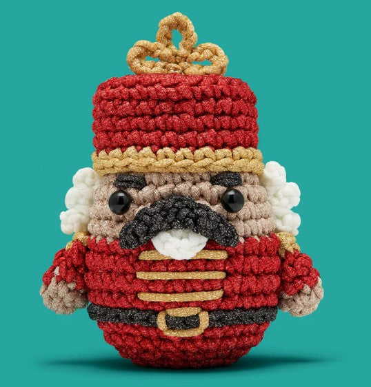 Hazel the Nutcracker Crochet Kit by The Woobles features a red hat and uniform, gold accents, black mustache, white curly hair, and black button eyes—perfect for step-by-step video lessons, set against a teal background.