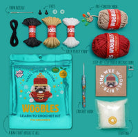 A flat lay of The Woobles Hazel the Nutcracker Crochet Kit, including yarn bundles, crochet hook, stuffing, illustrated instructions, plastic eyes, pre-started yarn, a teal pouch, and step-by-step video lessons for beginners.