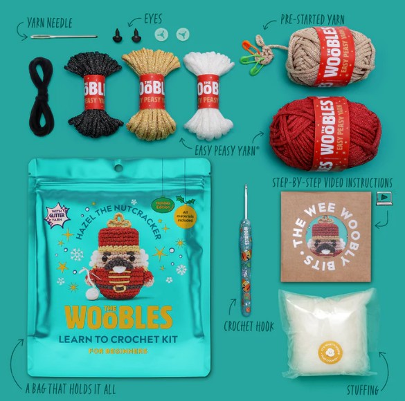A flat lay of The Woobles Hazel the Nutcracker Crochet Kit, including yarn bundles, crochet hook, stuffing, illustrated instructions, plastic eyes, pre-started yarn, a teal pouch, and step-by-step video lessons for beginners.