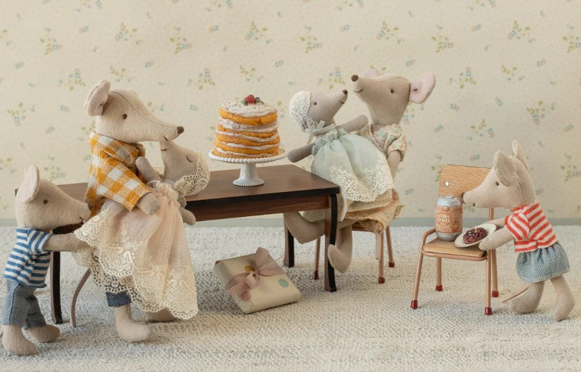 Four Maileg toy mice in clothes gather by a cake, as Maileg Baby Mouse In Gown - Mint sits at a tiny chair and table with a snack and drink. A wrapped gift from Maileg rests on the floor.