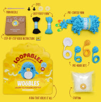 A flat lay of The Woobles Pierre the Penguin Loopables Kit displays a bag, yarn needle, eyes, yellow and blue yarn, instructions, pre-made yarn bits, stuffing, and pouch—ideal for beginner crafters.
