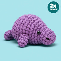 Phyllis the Manatee Extra Squeezy Crochet Kit by Woobles, a plush purple manatee toy, sits on a light blue background. A circular label in the corner says "2x BIGGER" in white text.