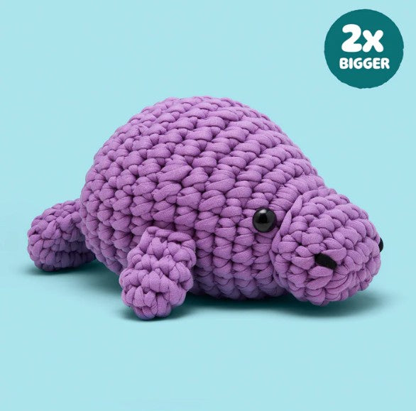 Phyllis the Manatee Extra Squeezy Crochet Kit by Woobles, a plush purple manatee toy, sits on a light blue background. A circular label in the corner says "2x BIGGER" in white text.