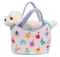 The Douglas Charmingly Yours Sassy Pet Sak features a plush white dog with pastel fur sitting in a light pink carry bag adorned with vibrant charms—like a smiley face, strawberry, rainbow, ice cream, butterfly, and flowers—for imaginative play.