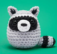 Scooter the Raccoon Crochet Kit by The Woobles features a plush amigurumi raccoon with black button eyes, a black mask, and a striped tail on a green background—ideal for beginner crochet projects or any crochet kit collection.